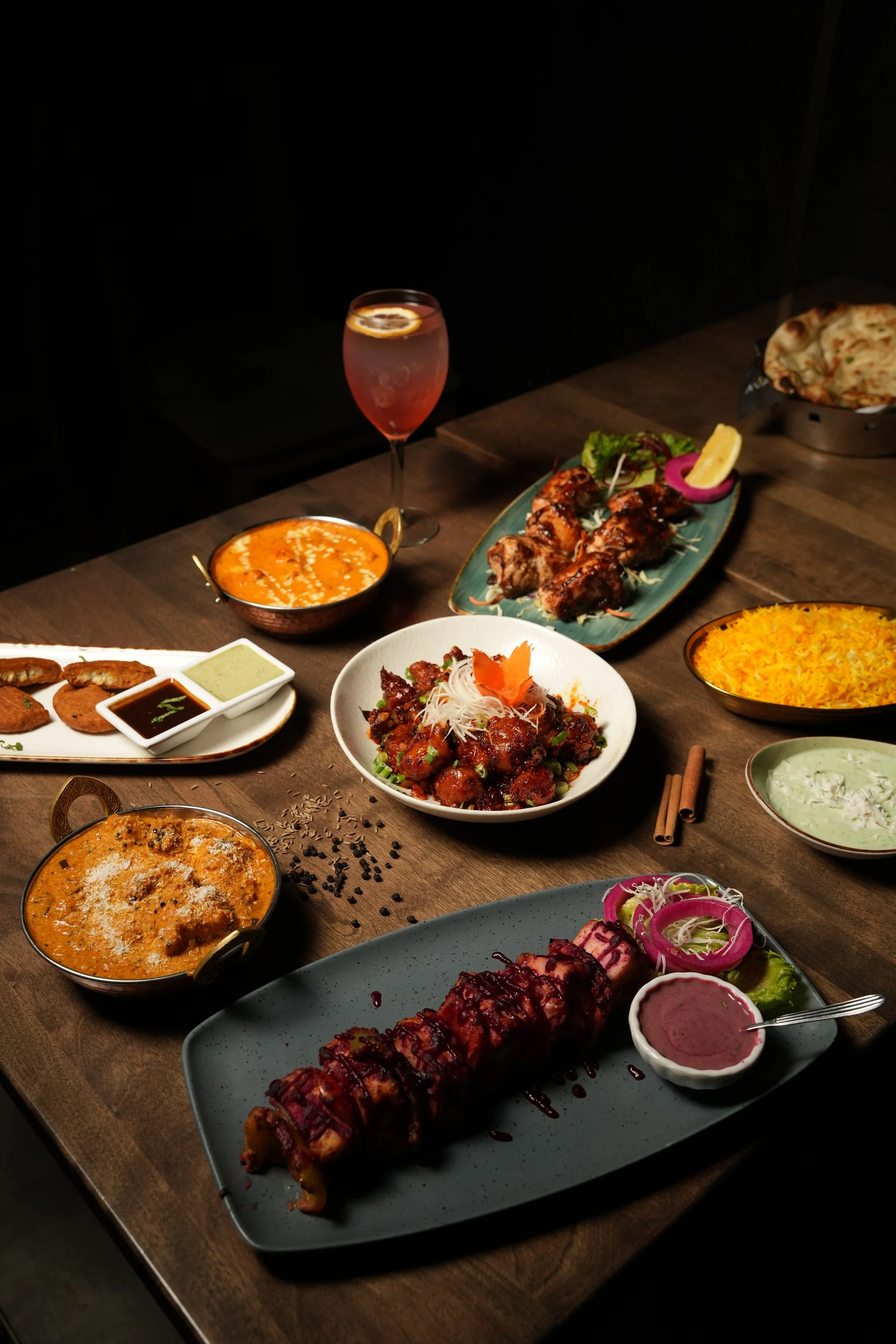 A vibrant spread featuring Sweet Potato Tikka, Mangalore Chicken Curry, Butter Chicken, Gobi 65, Maple Glazed Salmon, Garlic Naan, Saffron Rice, Pandan Pista Kheer, and our signature Cranberry Paneer Tikka. A balanced showcase of bold spices, comfort