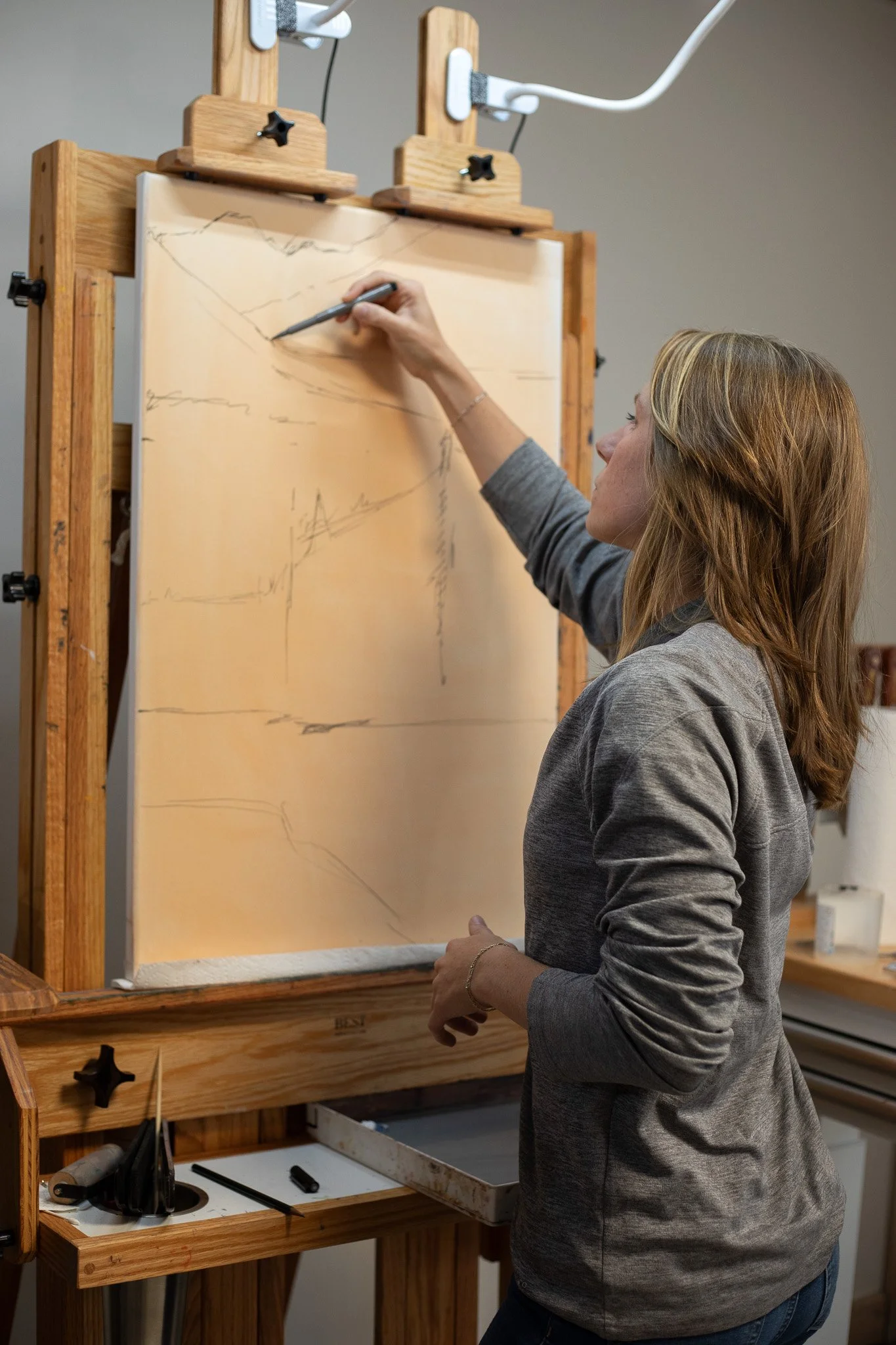 A woman drawing on a large canvas with a black marker in a wooden artist's easel. The canvas has sketch outlines. The artist's tools and supplies are on a workspace underneath.