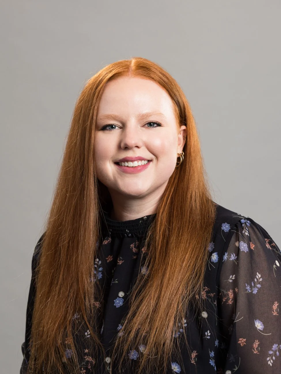 Portrait of a woman with long red hair smiling against a gray background, wearing a black floral blouse.