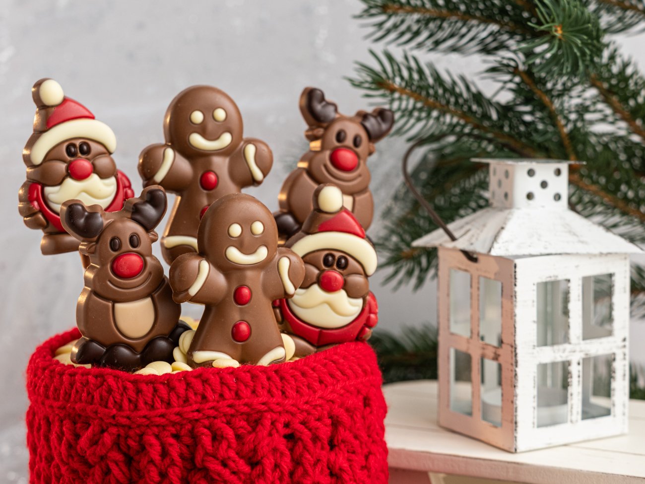 Chocolate Christmas chocolates featuring Santa Claus, gingerbread men, reindeer, and snowman in a red knitted container with a Christmas tree and white lantern in the background.