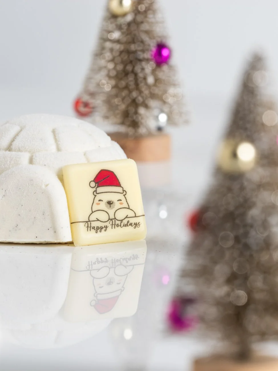 A white snow-like dome with a decorative white chocolate piece featuring a cartoon bear wearing a Santa hat, with the text "Happy Holidays" underneath, set against a blurred background of small Christmas trees with ornaments.