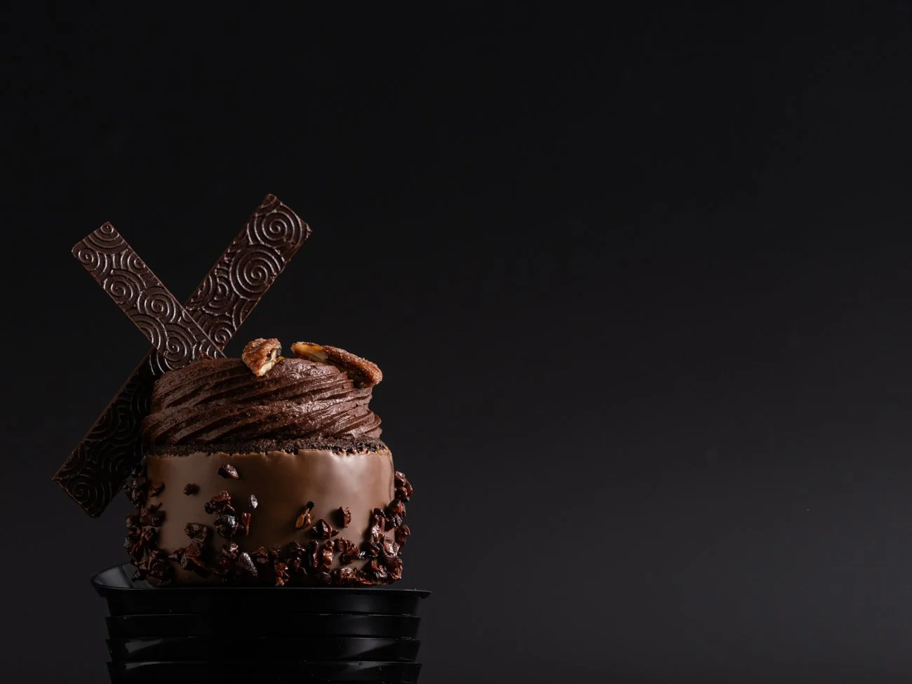 A chocolate dessert with dark chocolate, chocolate shavings, and decorative chocolate pieces on a black container against a black background.