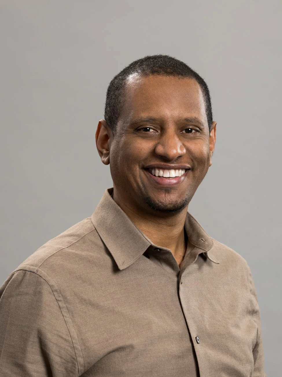 Portrait of a smiling man wearing a tan button-up shirt, standing against a plain grey background.