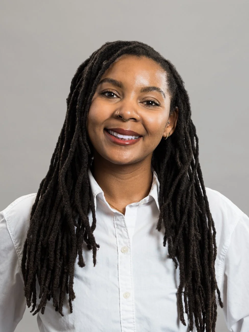 A woman with long dreadlocks, smiling, wearing a white collared shirt, posing against a gray background.