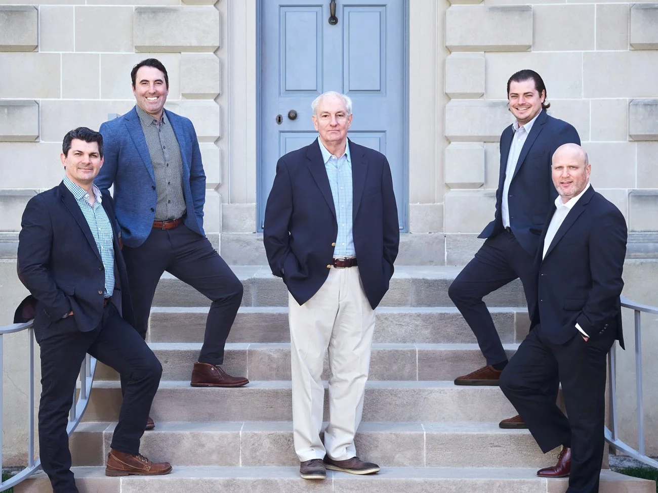 Group of six men in suits standing on steps in front of a stone building with a blue door