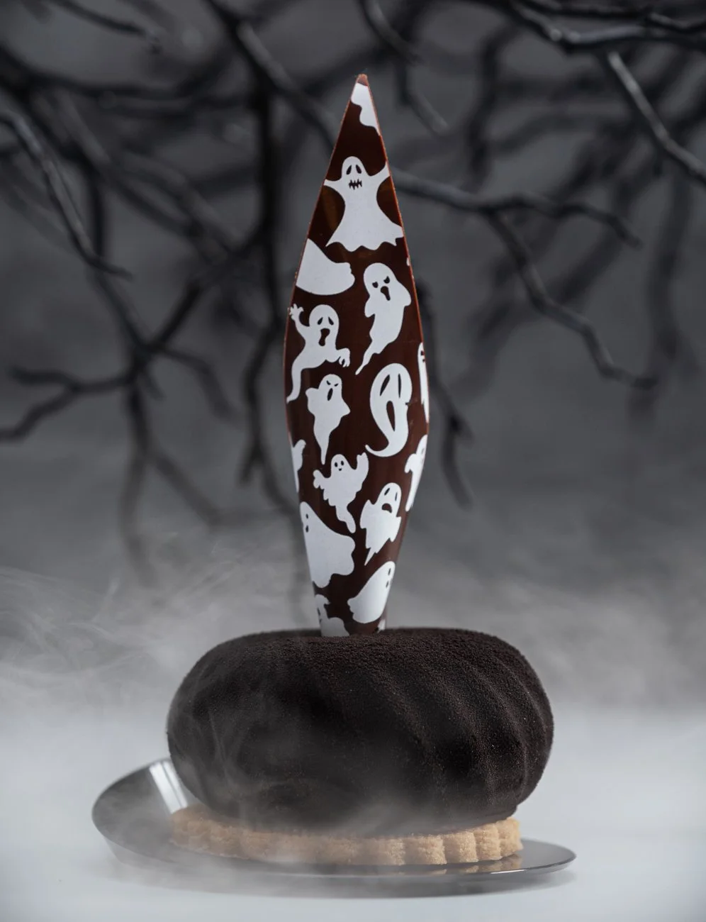 A decorative knife with a ghost pattern, displayed on a plate with a black stone and a cookie, with a spooky, smoky background and dark, twisted tree branches.