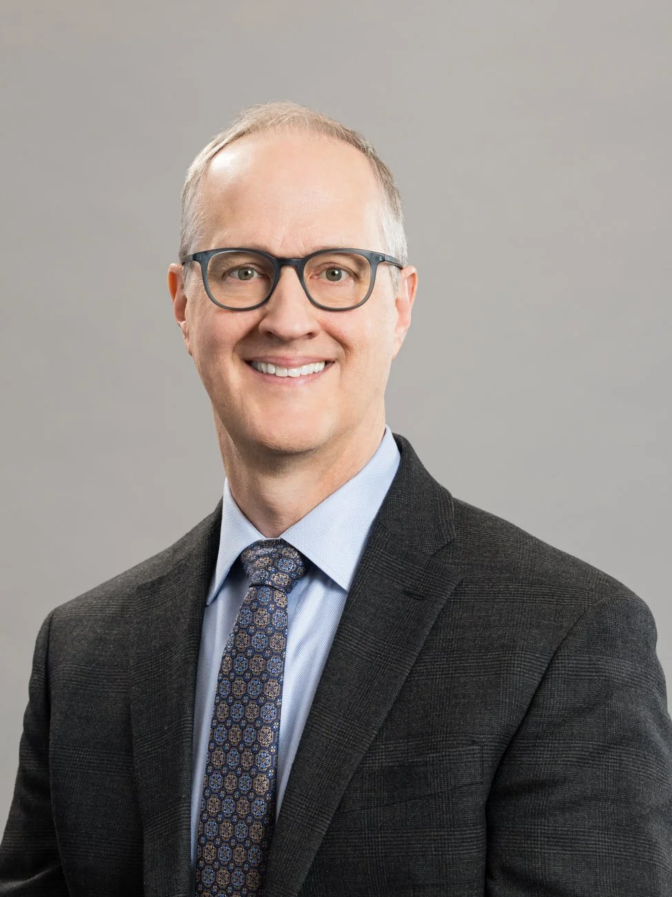 A professional portrait of a man wearing glasses, a dark suit, light blue shirt, and patterned tie, smiling against a gray background.