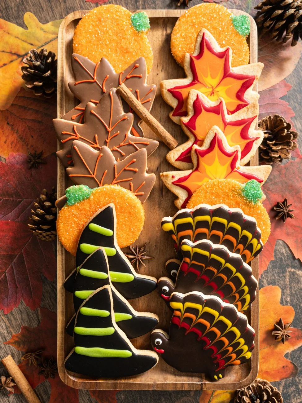Decorated autumn-themed cookies on a wooden tray, including maple leaves, acorns, a Halloween spider, and turkeys, with fall leaves, pinecones, star anise, and cinnamon sticks around.