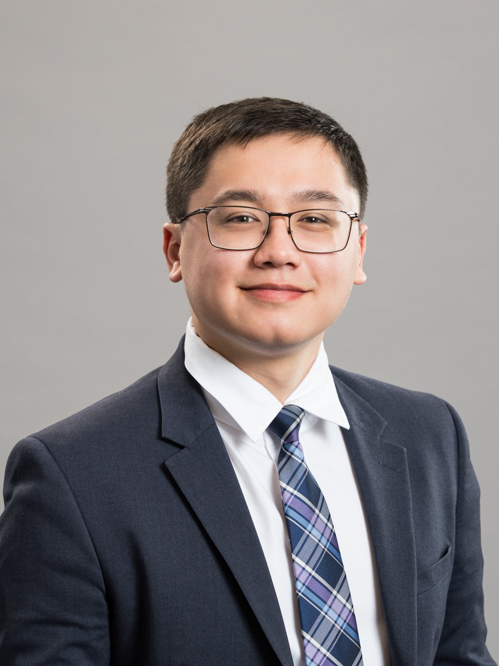 Professional portrait of a young man wearing glasses, a dark suit, white shirt, and a plaid tie, smiling against a neutral gray background.