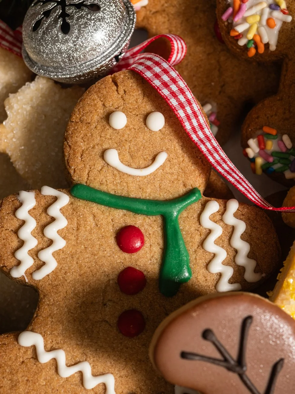 Close-up of a decorated gingerbread cookie with a smiling face, green icing scarf, and red buttons, surrounded by other decorated cookies and Christmas ornaments.