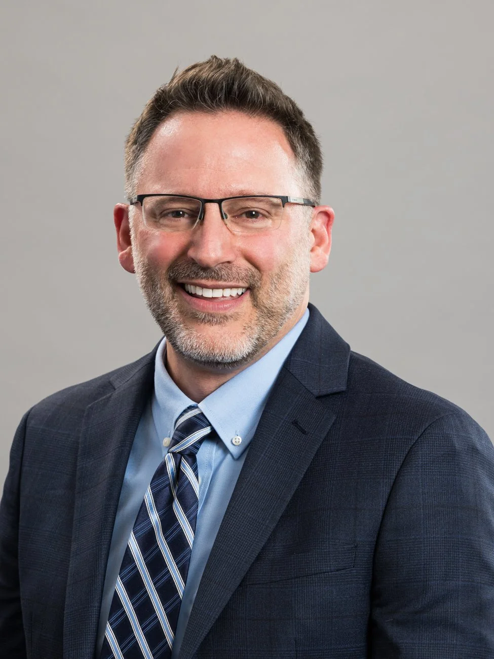 Headshot of a smiling middle-aged man with glasses, wearing a dark suit, light blue shirt, and striped tie against a neutral background.