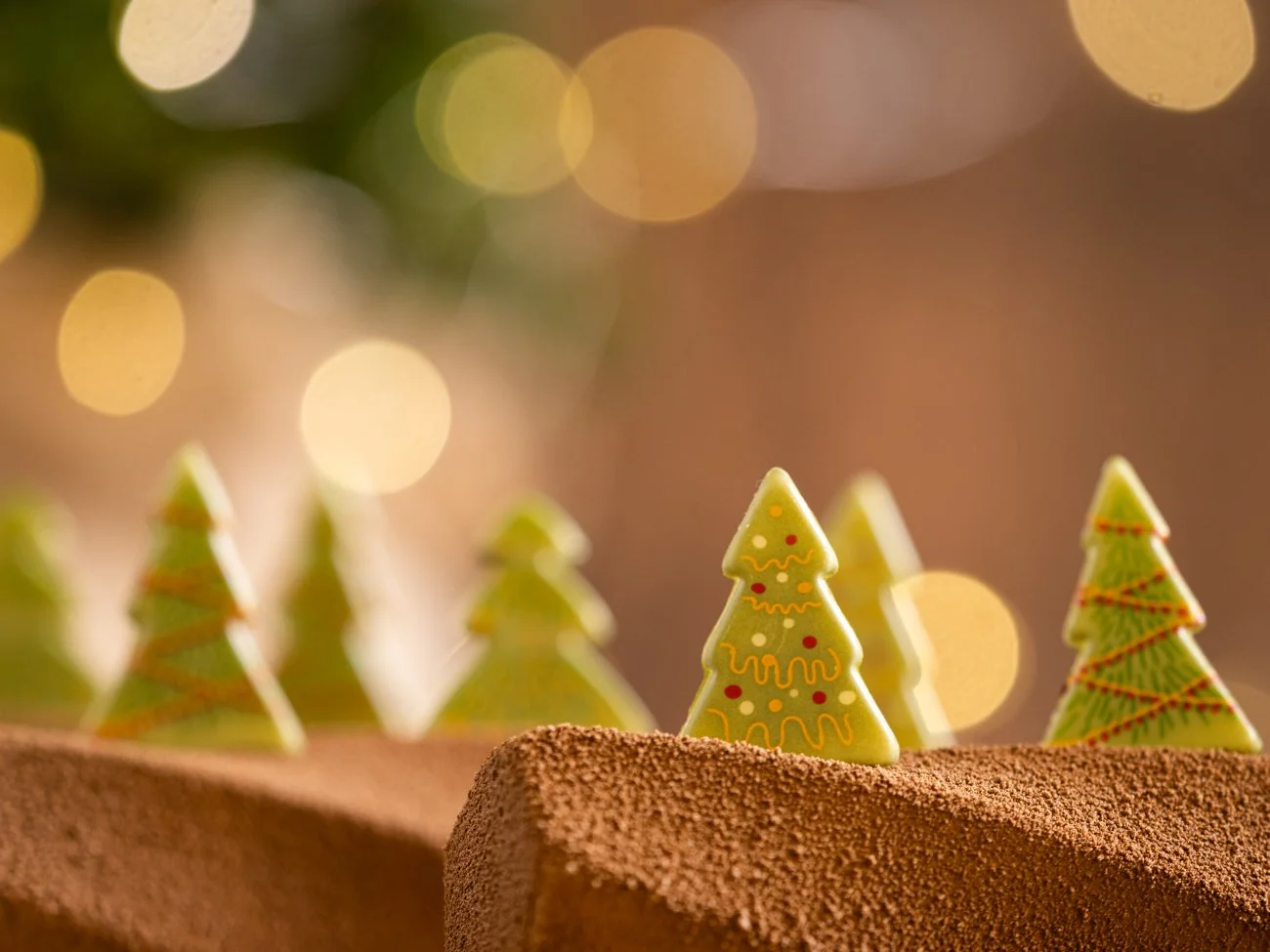 Decorative Christmas trees on a chocolate cake with a bokeh background of warm, glowing lights.