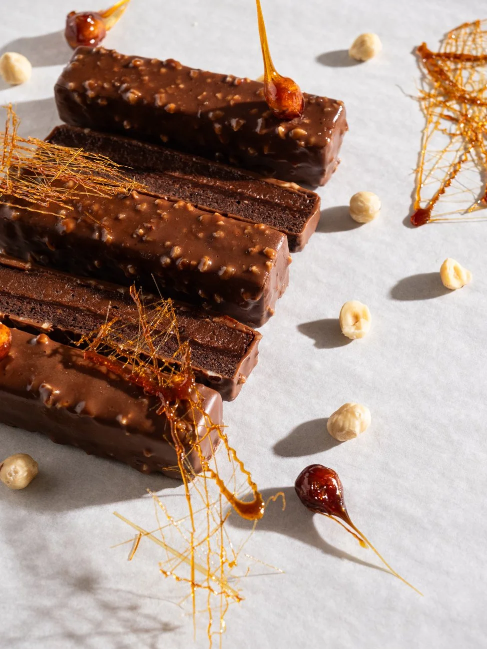 Close-up of three bars of chocolate with a glossy coating and texture, garnished with caramel decorations and scattered white and brown small round candies on a white surface.