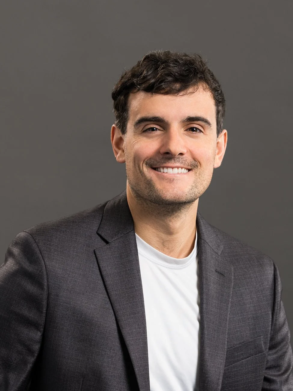 A man in a dark gray blazer and white shirt smiling against a plain gray background.