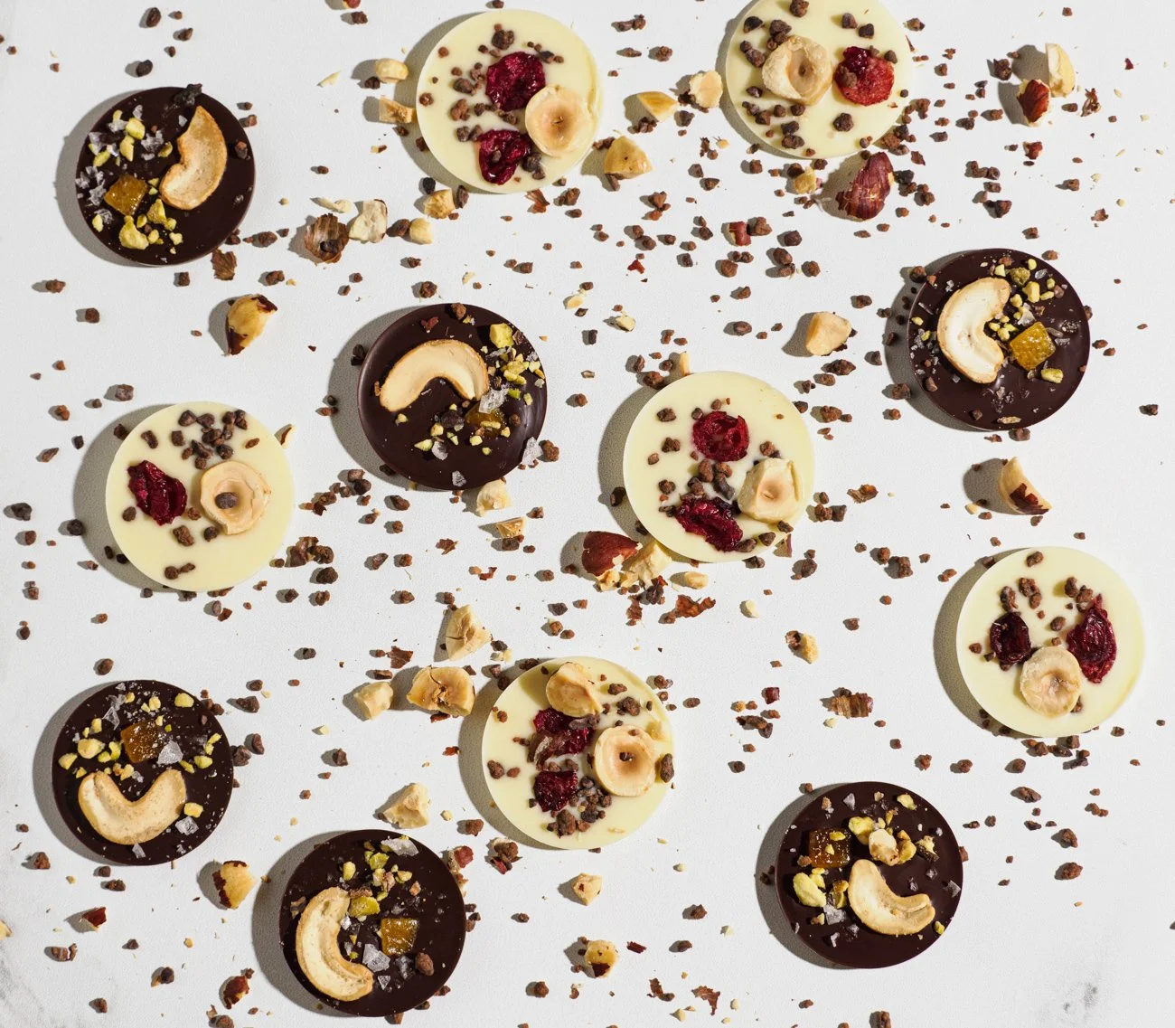 Assorted white and dark chocolates topped with nuts and dried fruit, scattered on a white surface.