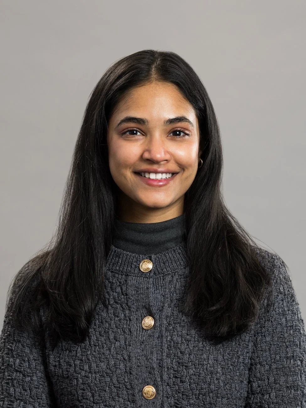 A woman with long black hair smiling at the camera, wearing a dark turtleneck and a gray buttoned cardigan, standing against a plain gray background.