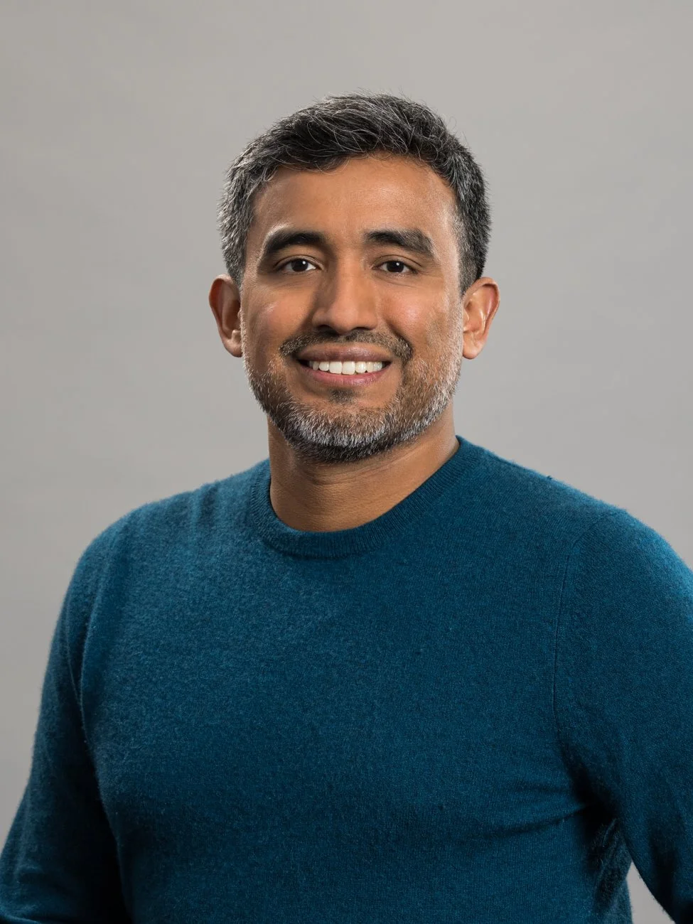 Headshot of a smiling man with short dark hair and a beard, wearing a teal sweater, standing against a gray background.