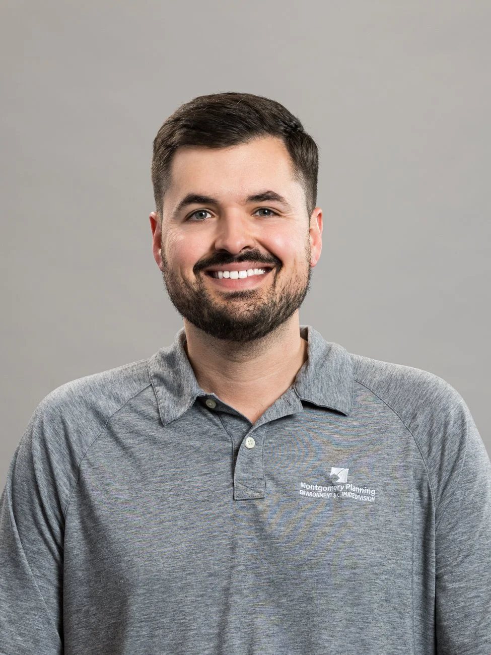 Portrait of a smiling man with short dark hair and a beard, wearing a gray polo shirt with Montgomery Planning logo, against a plain gray background.