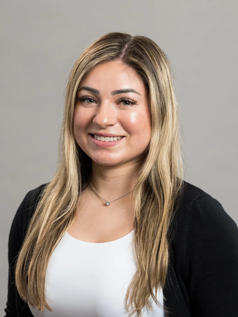 A professional young woman with long blond hair, wearing a white top, black cardigan, and a heart-shaped necklace, smiling against a plain gray background.