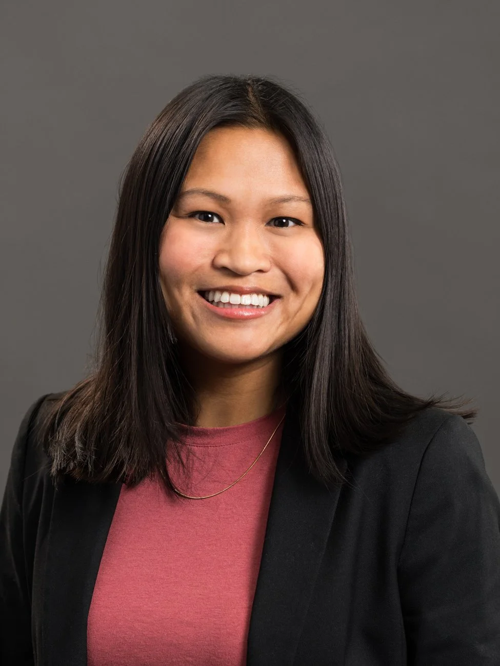 A portrait of a smiling woman with long black hair wearing a black blazer and a pink top against a gray background.