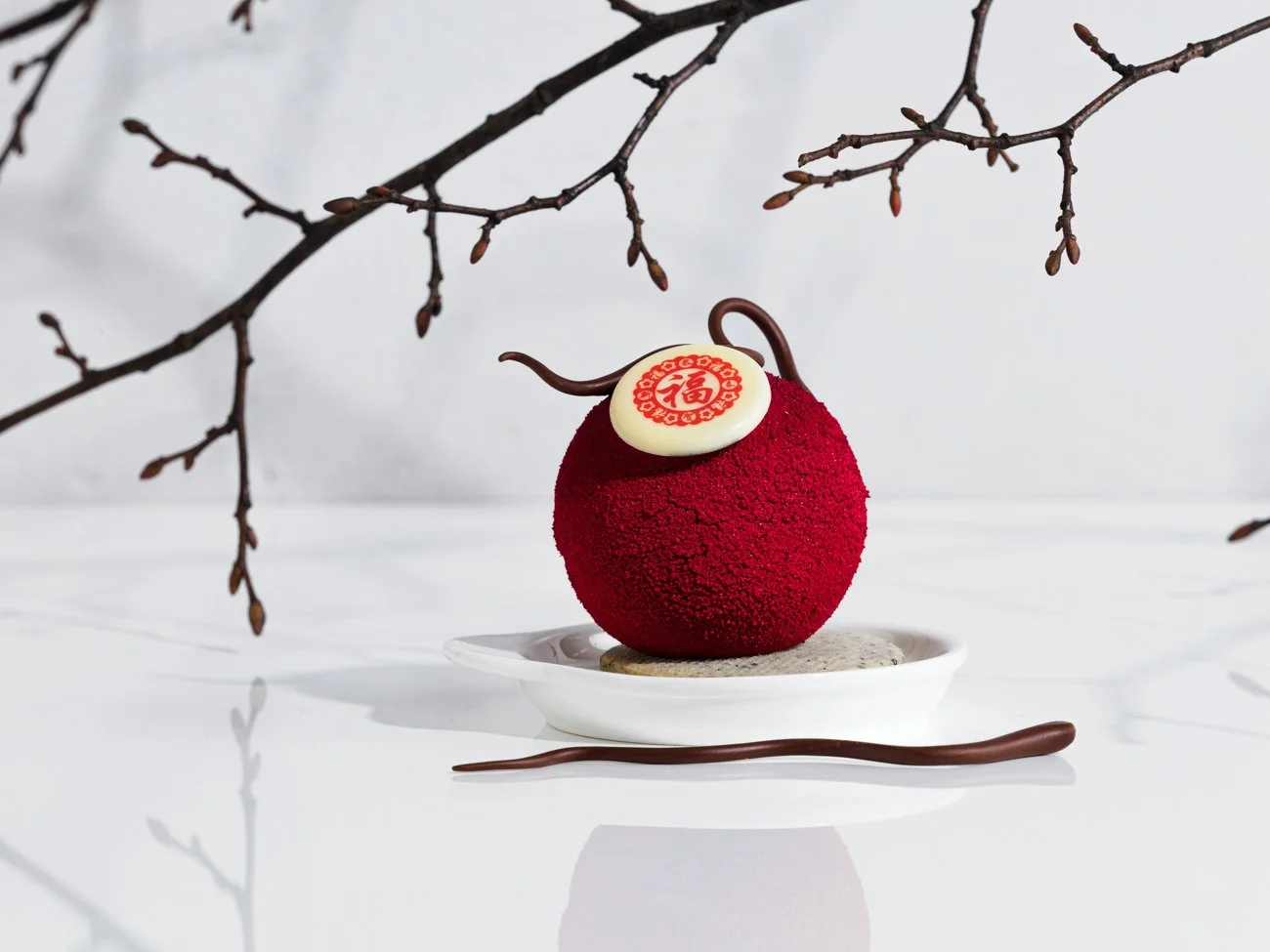 A decorative red and white cake shaped like a peach, with a white chocolate disc on top featuring a red Chinese character, on a white plate with branches overhead, on a white surface.