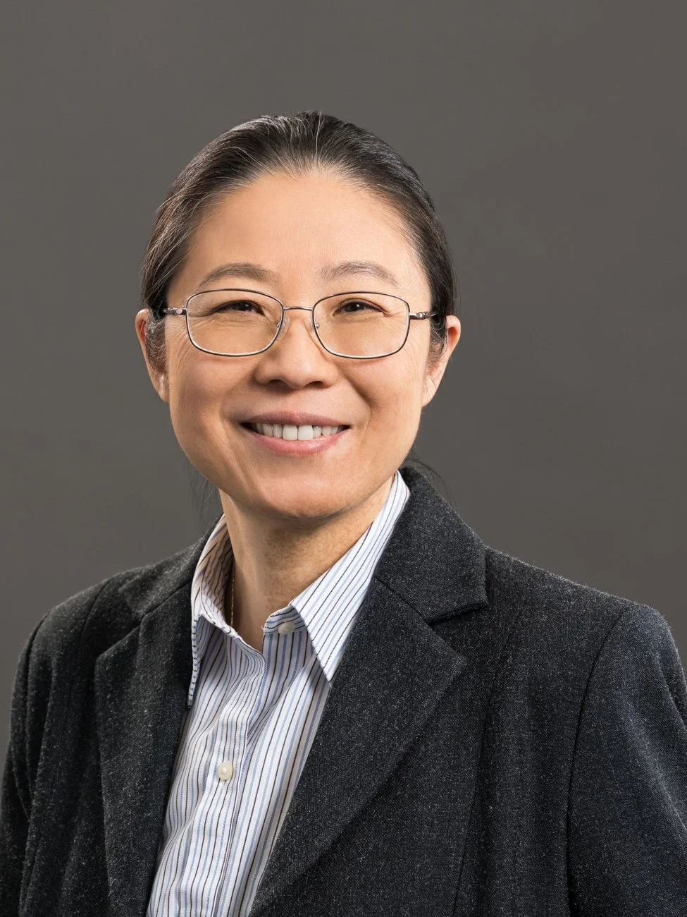 Professional woman with glasses smiling, wearing a dark blazer over a light striped shirt, against a gray background.