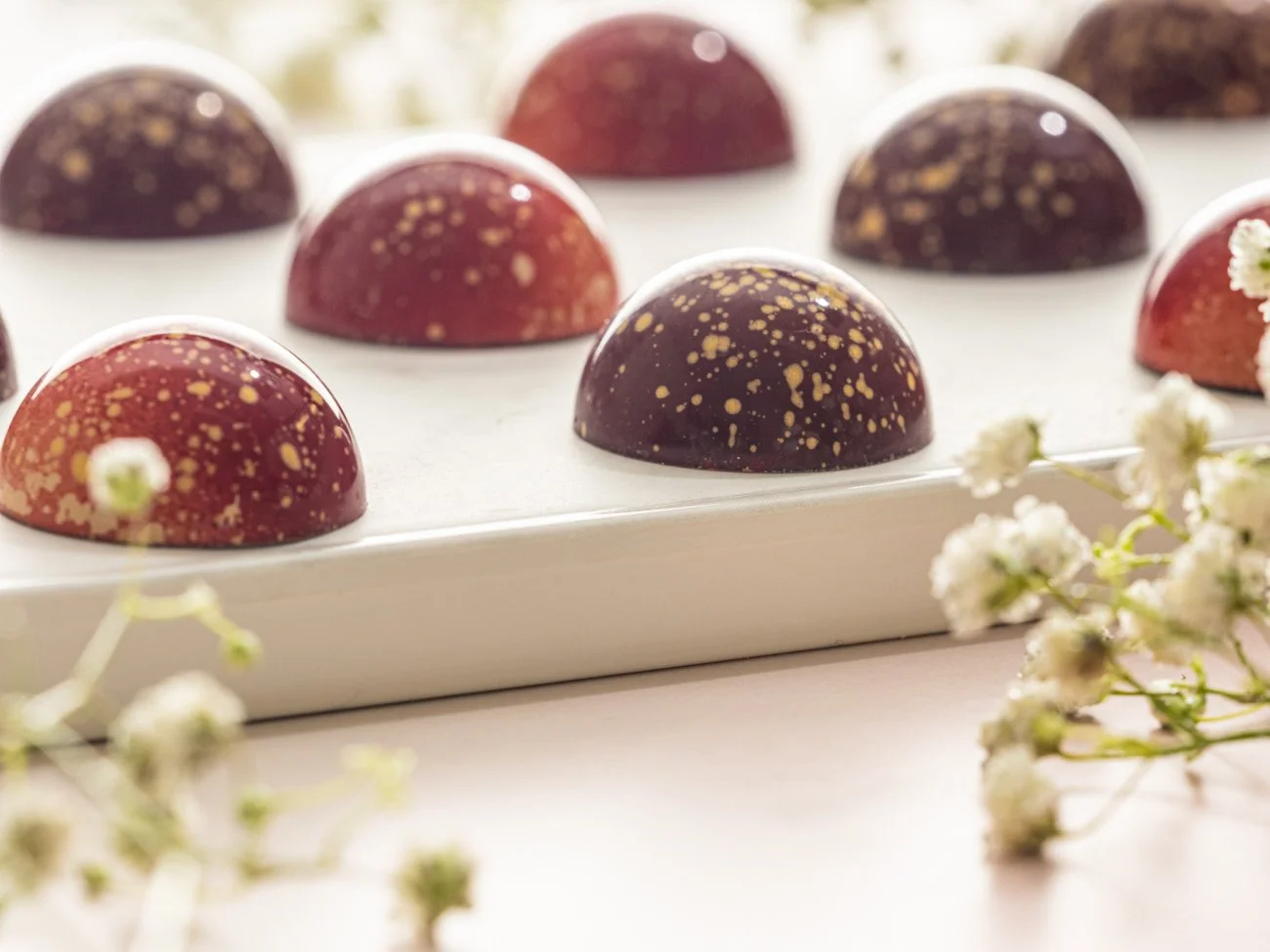 Chocolate truffles with gold splatter on a white platter, surrounded by small white flowers.
