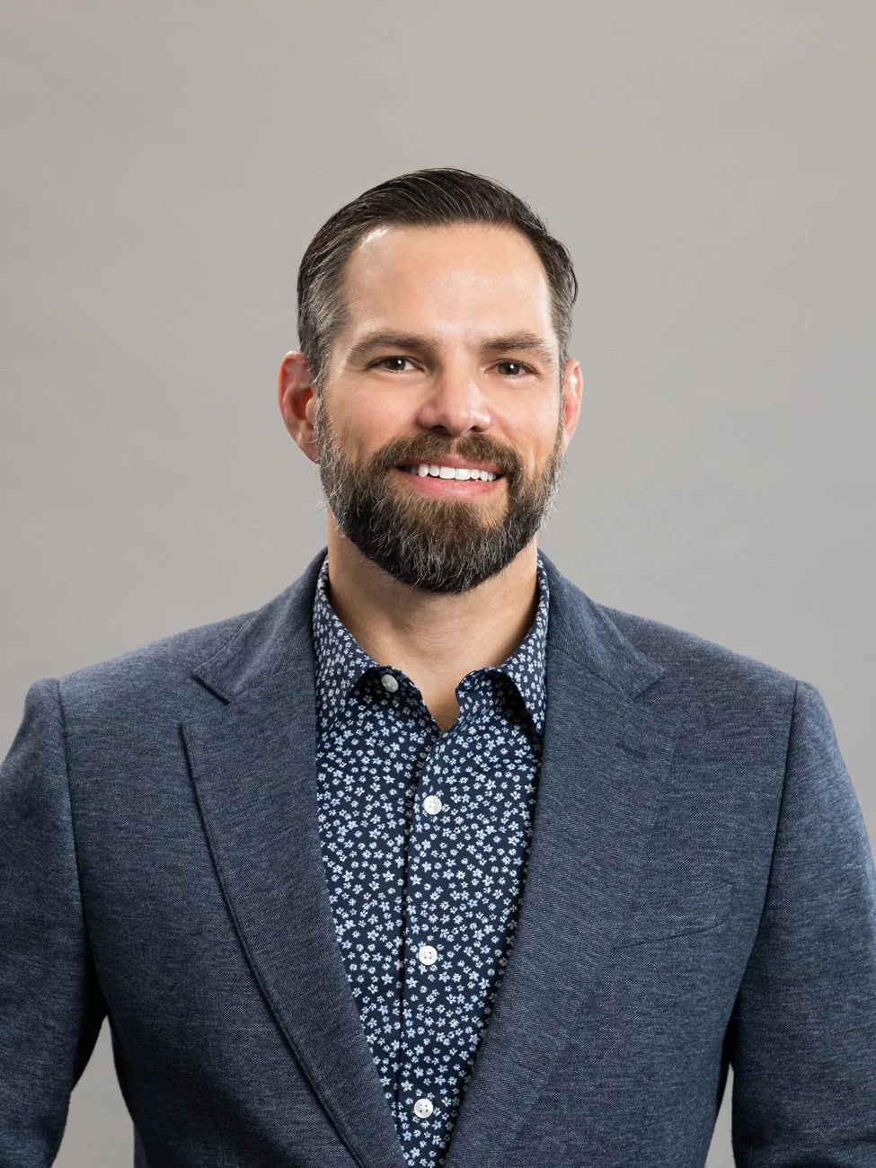 A professional man with dark hair, a beard, and a mustache, wearing a patterned dress shirt and a blazer, smiling against a plain background.