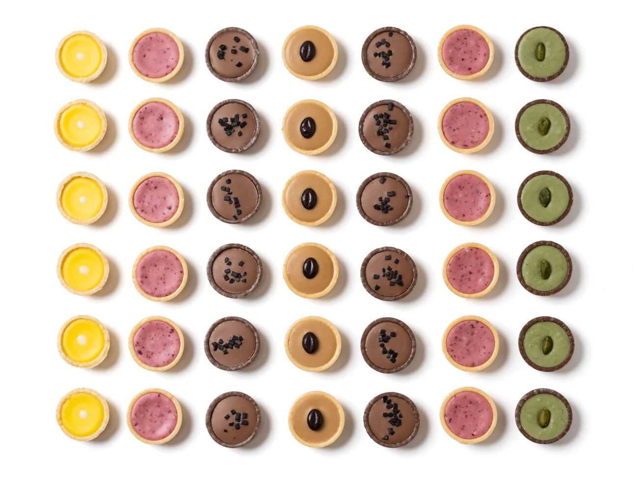 Assorted mini tarts with yellow, pink, chocolate, caramel, and green fillings, topped with small decorations, arranged in a grid on a white background.