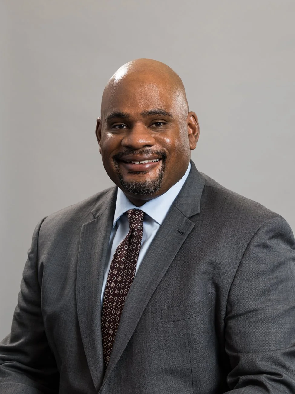 A smiling African American man in a gray suit and patterned tie, posing against a neutral gray background.