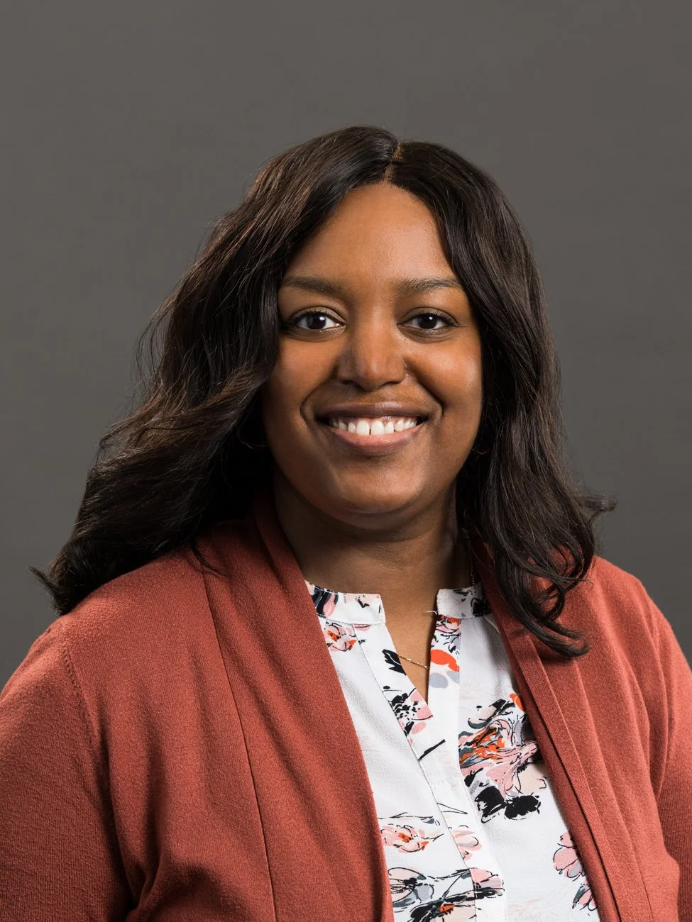 Portrait of a woman with dark hair smiling, wearing a burnt orange blazer and floral blouse against a gray background.