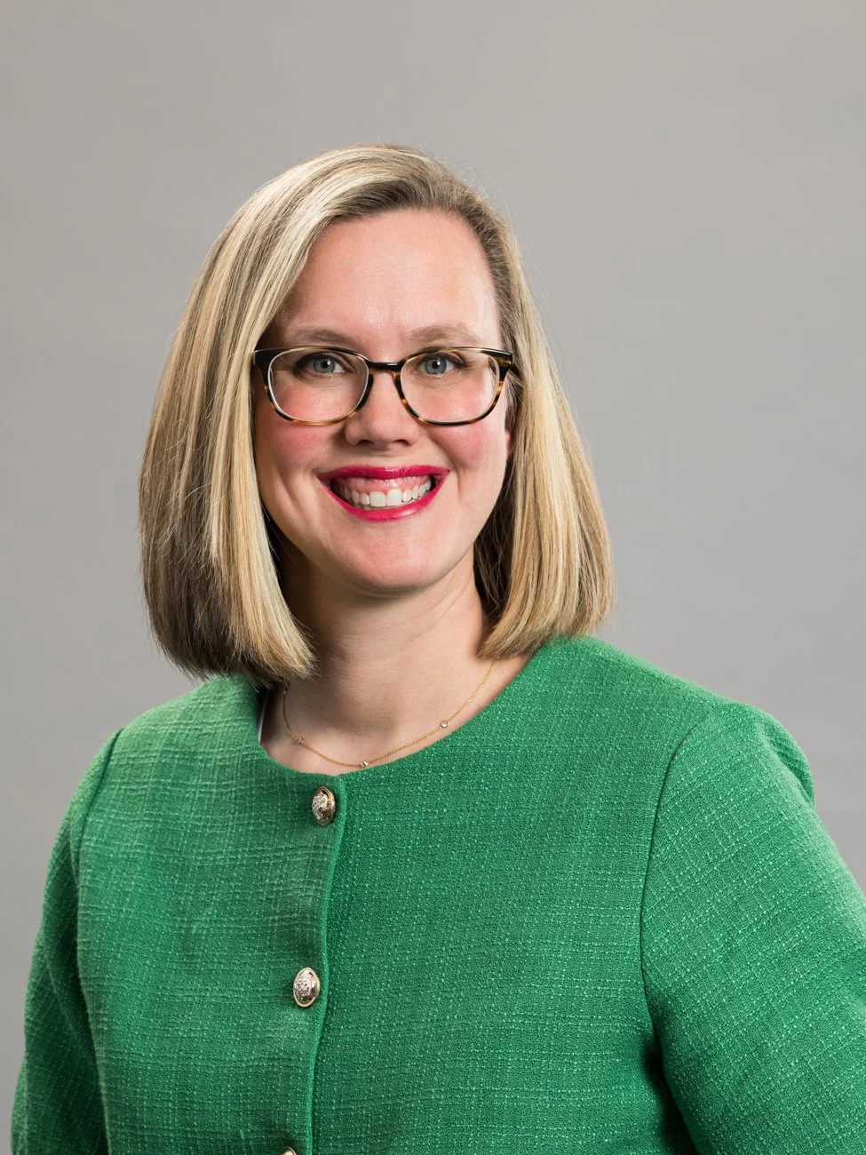 A woman with shoulder-length blonde hair, glasses, and a bright smile, wearing a green textured jacket with gold buttons, standing against a plain gray background.