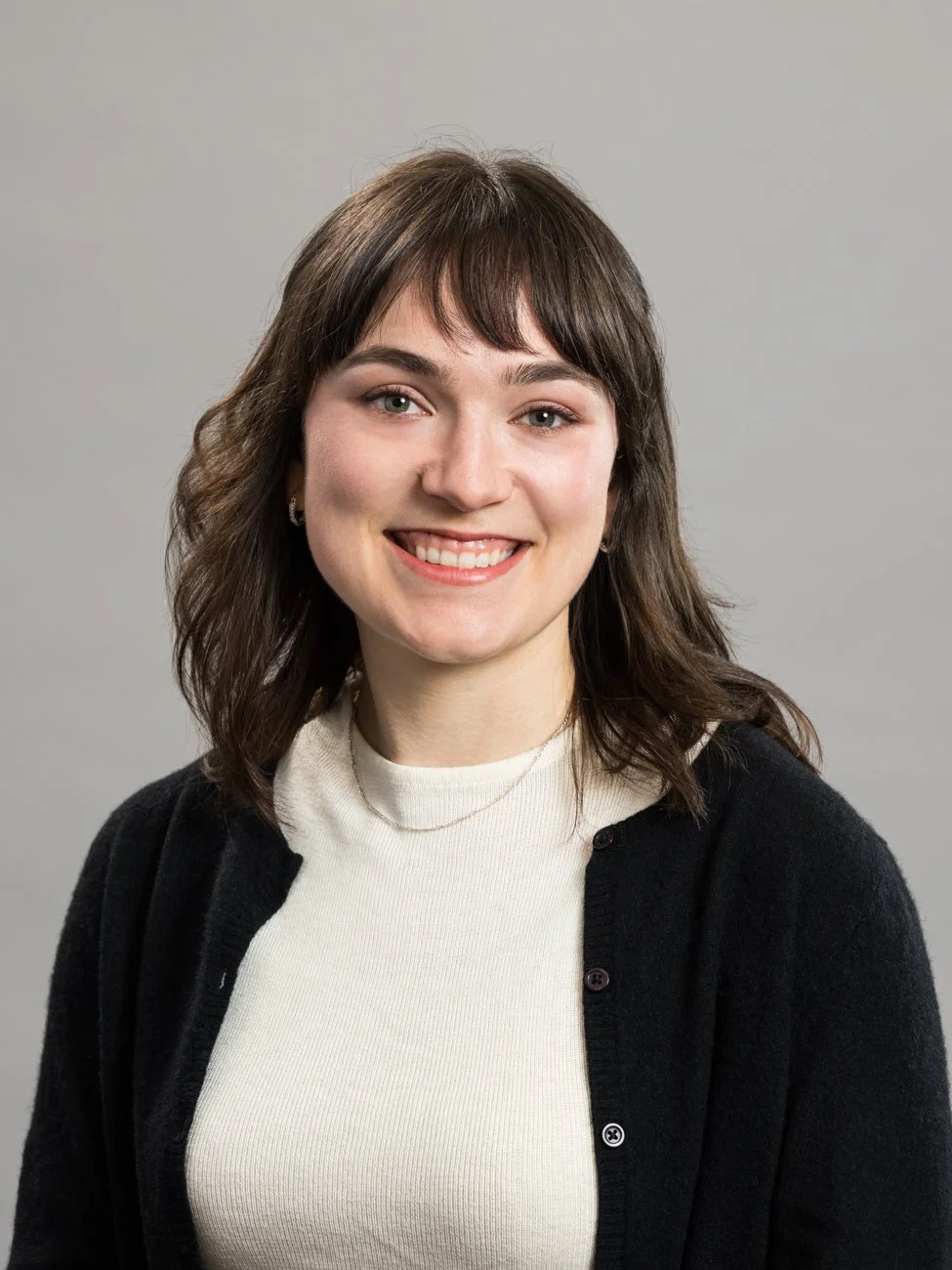 A young woman with shoulder-length brown hair and light skin, smiling in front of a plain gray background, wearing a white top and a black cardigan.