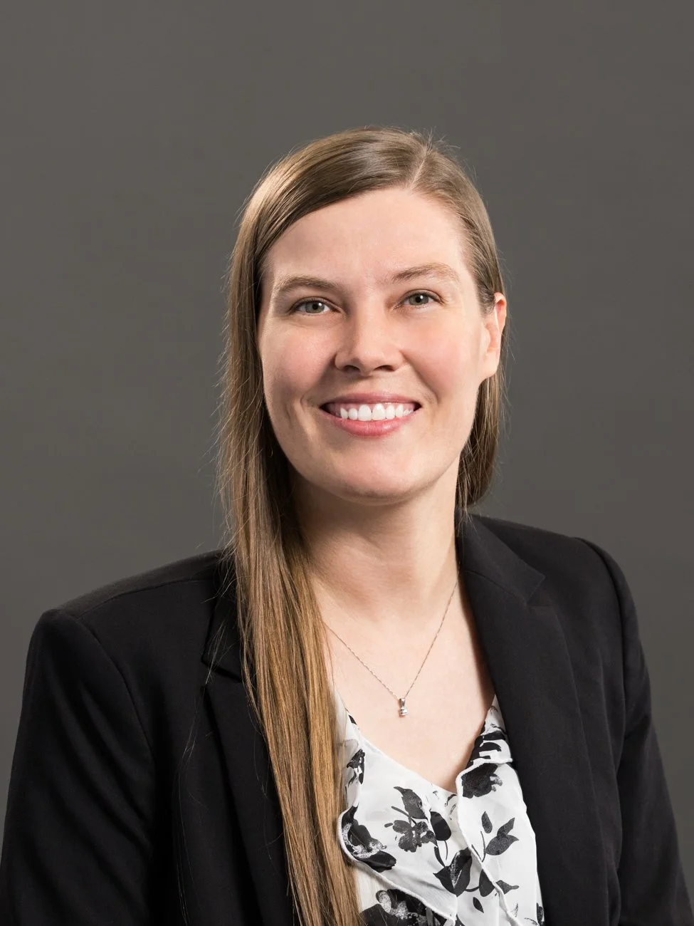 Professional portrait of a smiling woman with long brown hair, wearing a black blazer and a white blouse with black floral pattern, against a gray background.
