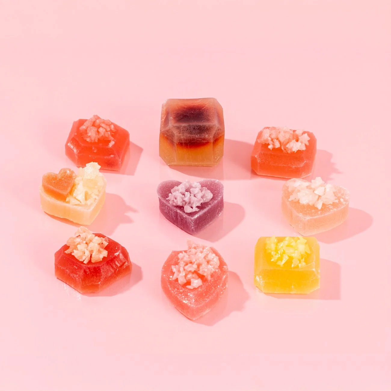 Assorted colorful heart-shaped and square-shaped candies with toppings on a pink background.