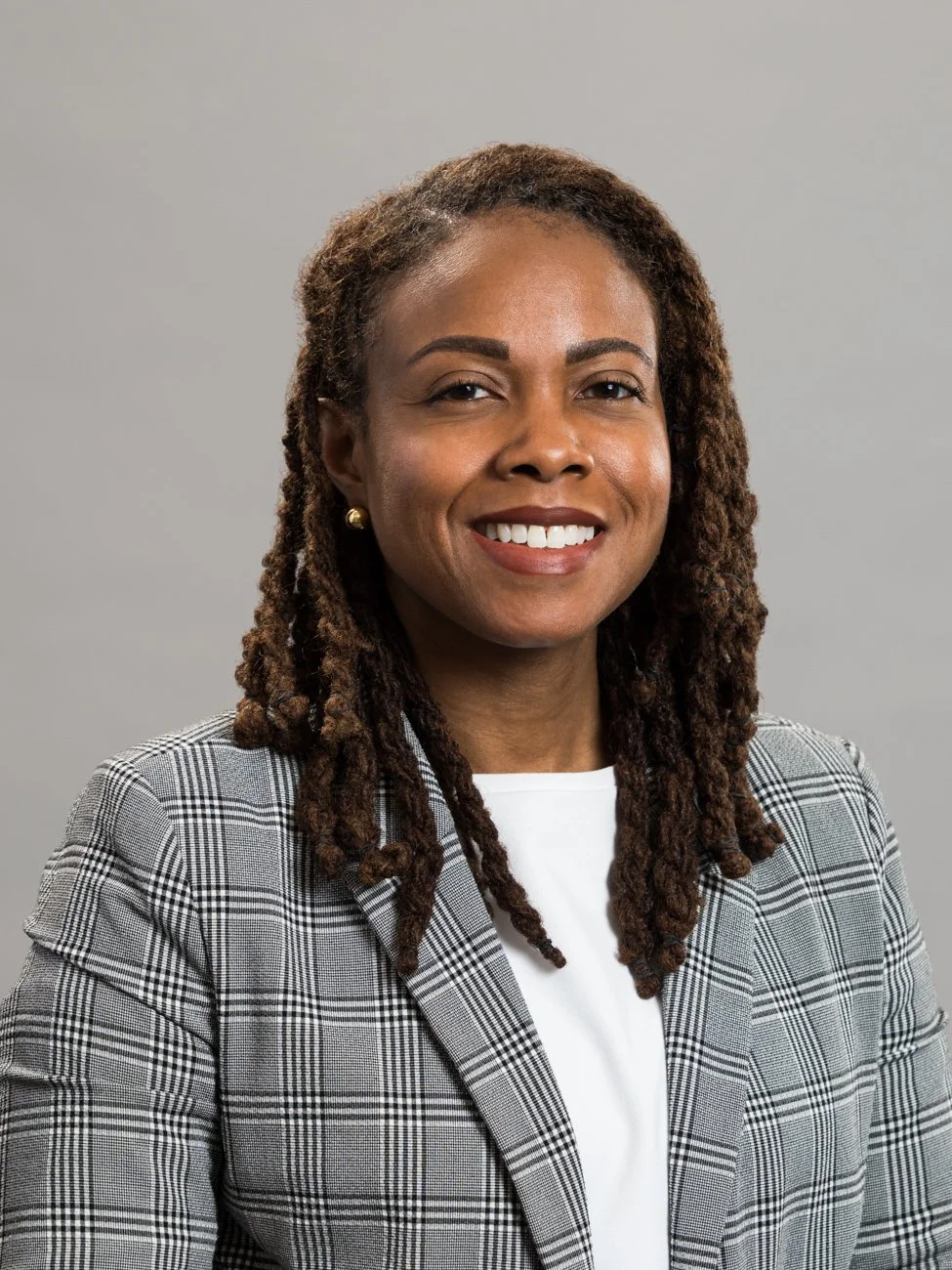 A professional woman with medium brown skin, locking brown hair, wearing a plaid blazer and smiling.