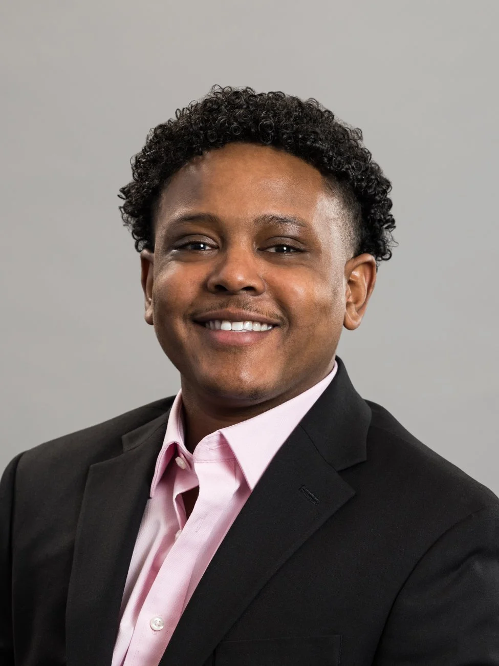 Headshot of a smiling man with short curly black hair wearing a black suit jacket and a light pink shirt against a plain gray background.