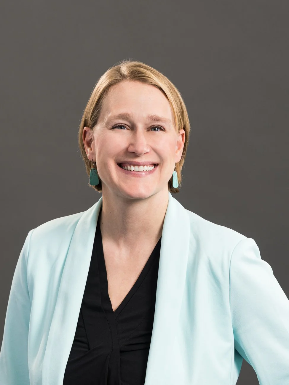 Portrait of a woman with short blonde hair wearing a light blue blazer, black top, and teal earrings, smiling against a gray background.