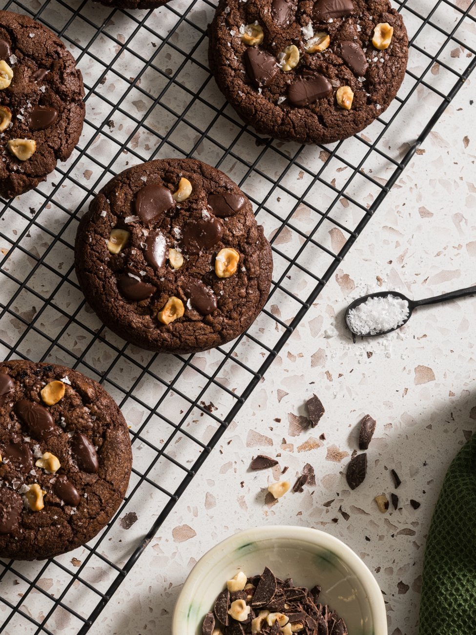 Chocolate cookies with chunks of chocolate and chopped nuts on a cooling rack, with a small bowl of chopped chocolate and nuts nearby.