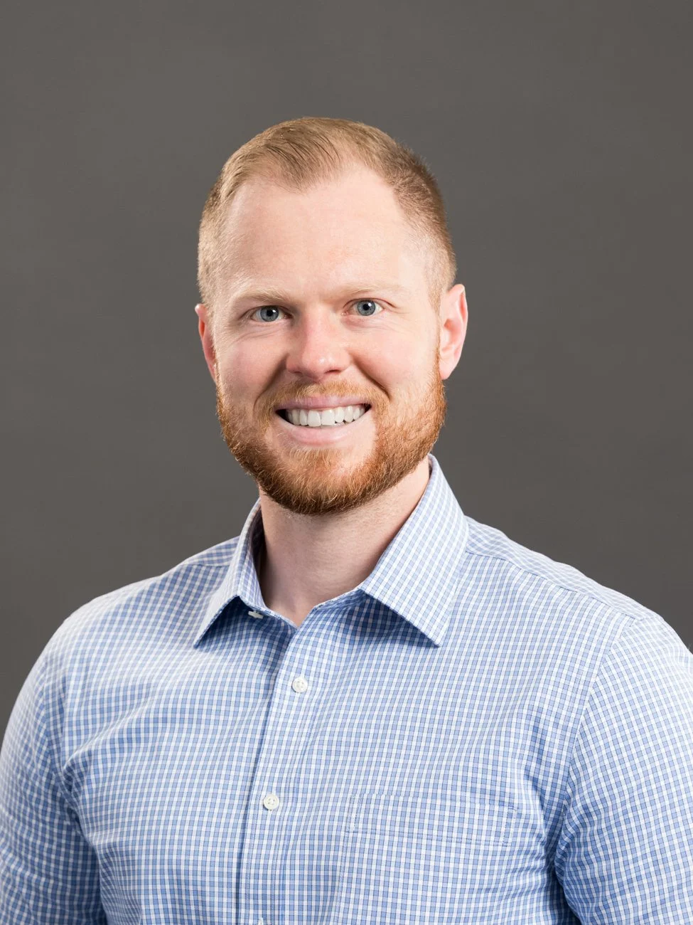 Headshot of a smiling man with red hair and beard, wearing a blue checkered shirt, against a gray background.