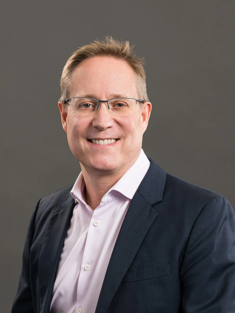 A smiling man in a suit with glasses, wearing a light pink shirt, against a plain gray background.