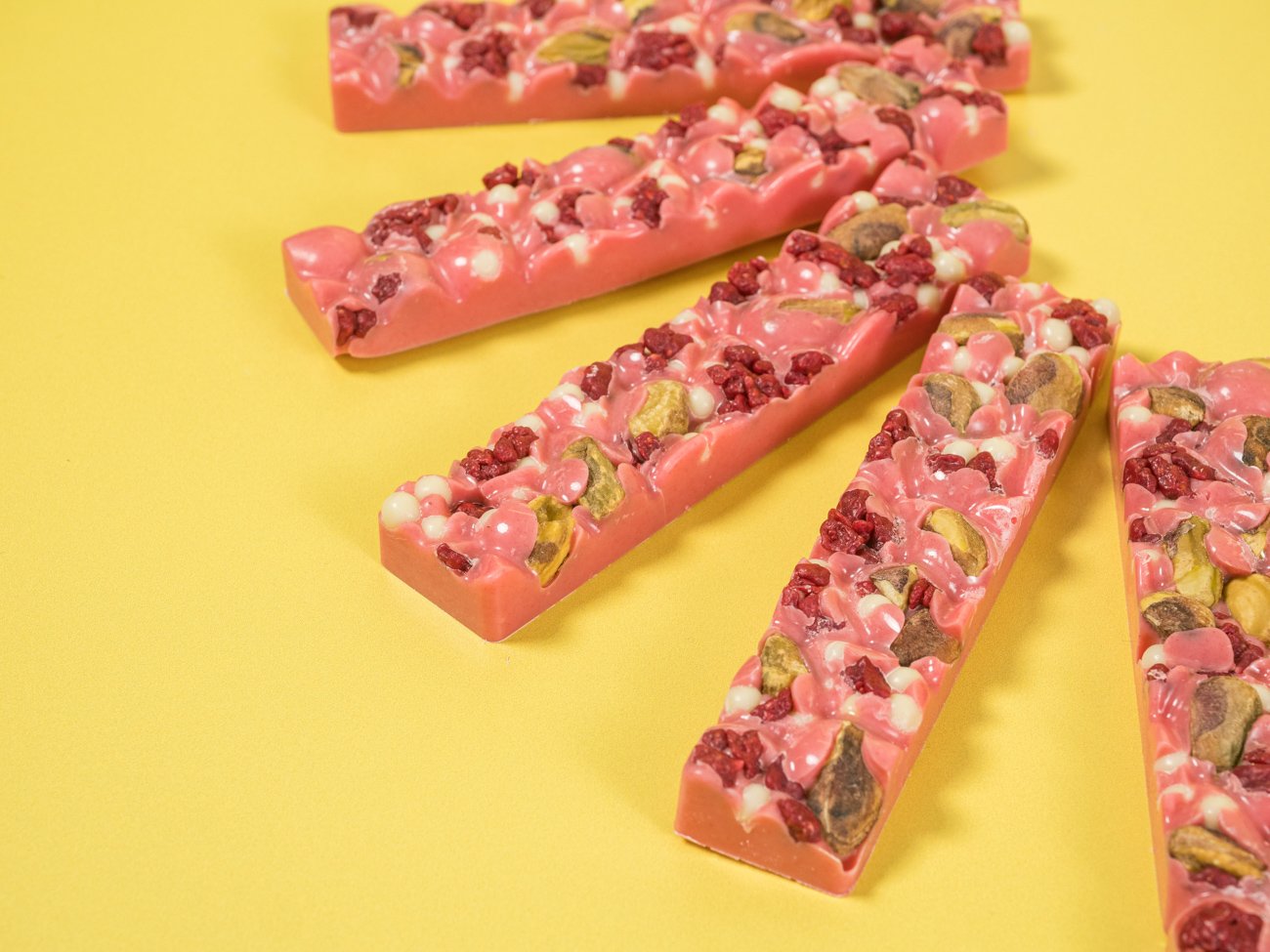 Close-up of pink chocolate bars embedded with pistachios, freeze-dried raspberries, and white chocolate chips on a yellow background.