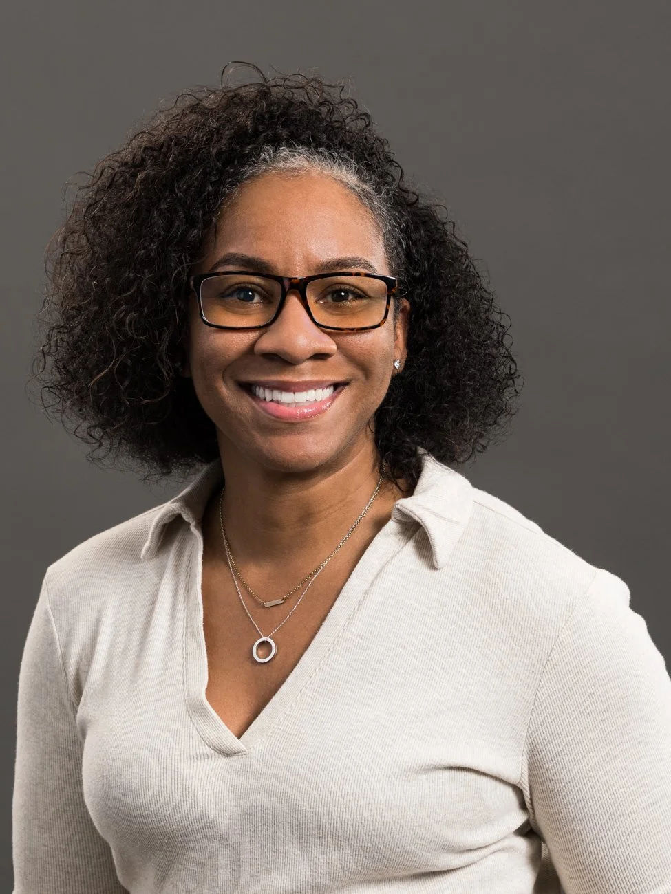 Portrait of an African American woman with curly hair, wearing glasses and a beige collared top, smiling against a gray background.