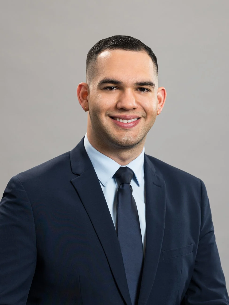 Professional portrait of a smiling man with short dark hair, wearing a navy blue suit, light blue dress shirt, and navy tie, against a plain gray background.