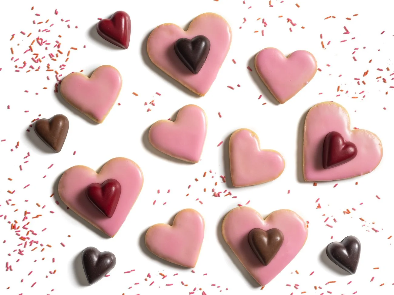 Heart-shaped pink cookies with small chocolate hearts on top, scattered among red and pink sprinkles on a white background.