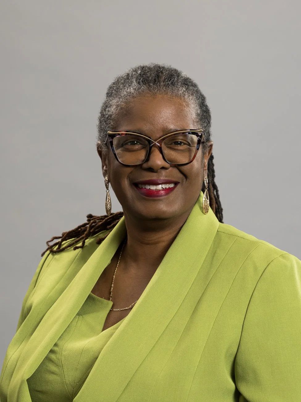 A woman with gray hair, wearing glasses and earrings, smiling, dressed in a bright green blazer and matching top, standing against a plain gray background.