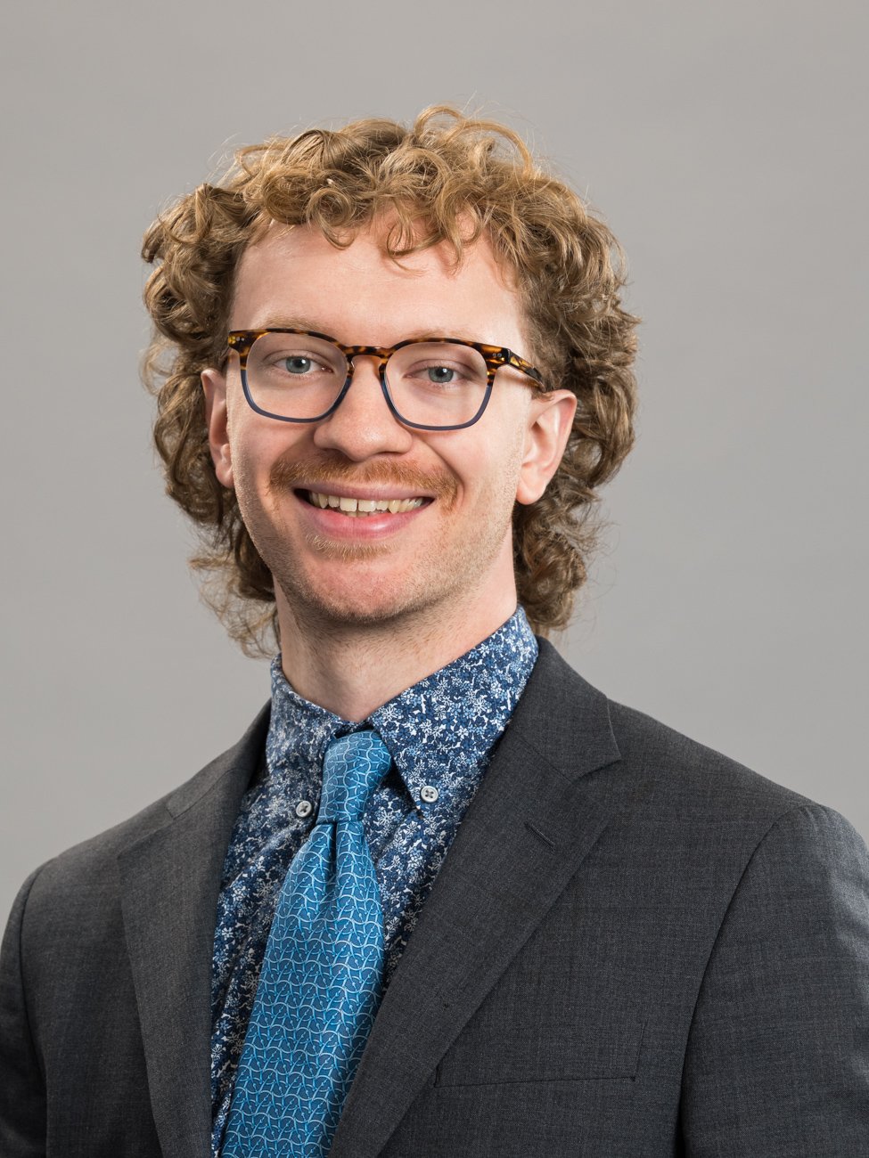 Portrait of a man with curly hair, wearing glasses, a patterned shirt, a blue tie, and a dark suit, smiling against a gray background.