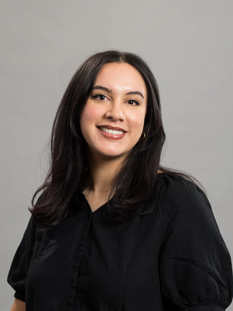 A woman with long dark hair, wearing a black shirt, smiling against a gray background.