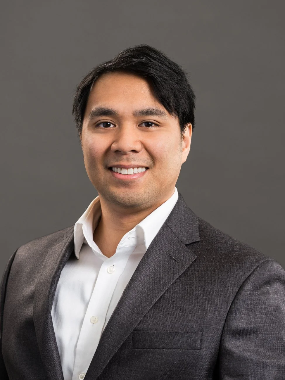 Professional headshot of a man with black hair, wearing a gray suit jacket and white dress shirt, smiling against a gray background.