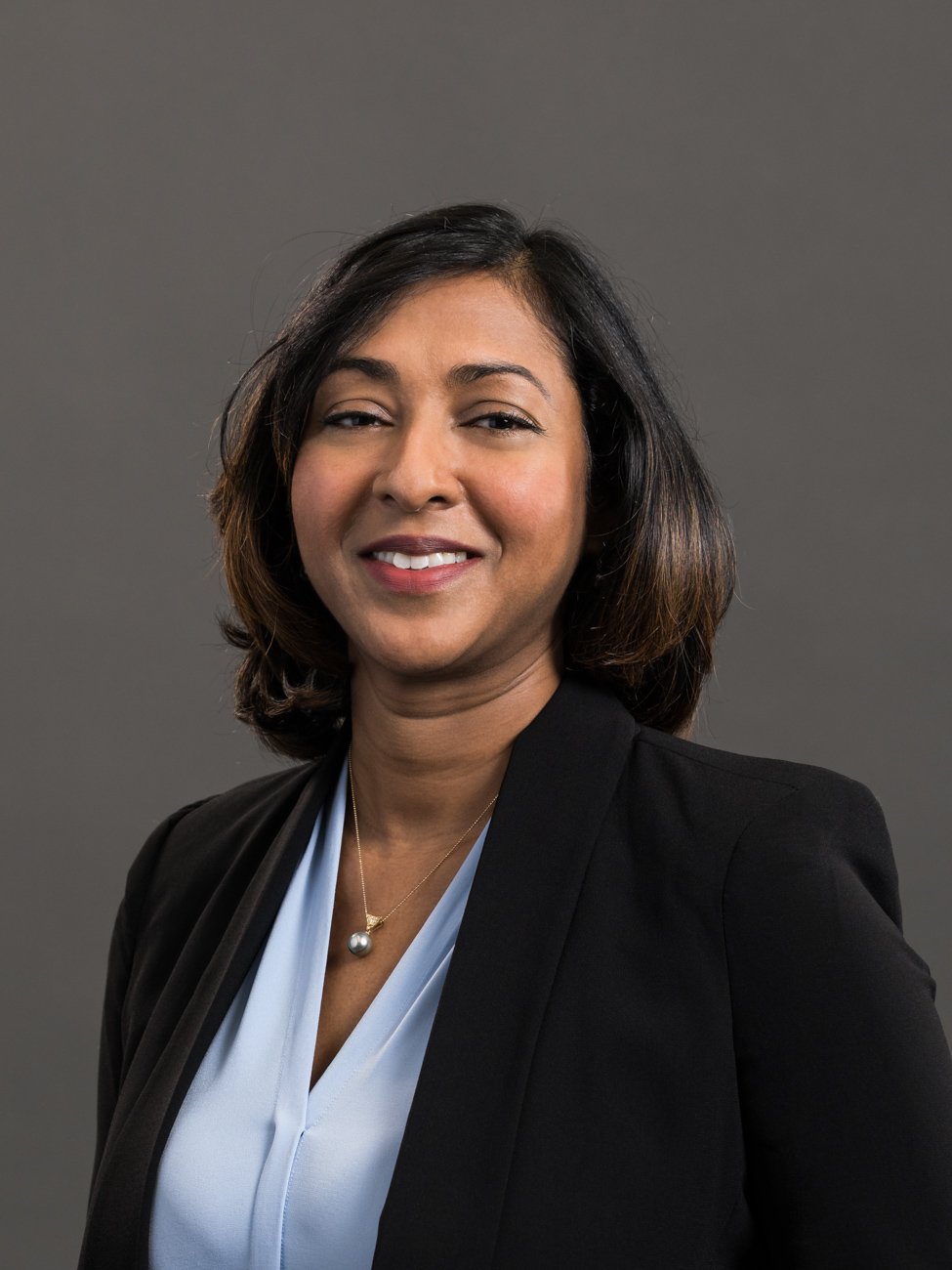 Professional woman with shoulder-length dark hair, wearing a black blazer, light blue blouse, and a pearl necklace, smiling against a gray background.